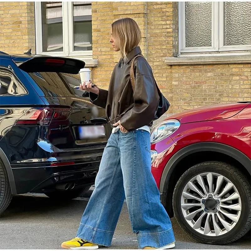 Stylish woman in oversized brown leather jacket, wide-leg jeans, and yellow sneakers walking outdoors.