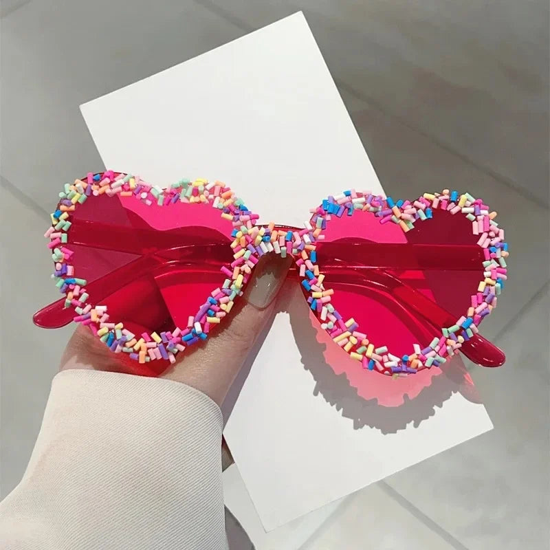 Heart-shaped pink sunglasses with rainbow sprinkle detailing, held by a hand
