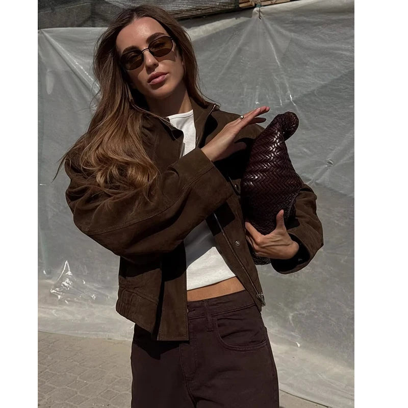 Woman in brown suede jacket and sunglasses holding a textured dark brown clutch bag.