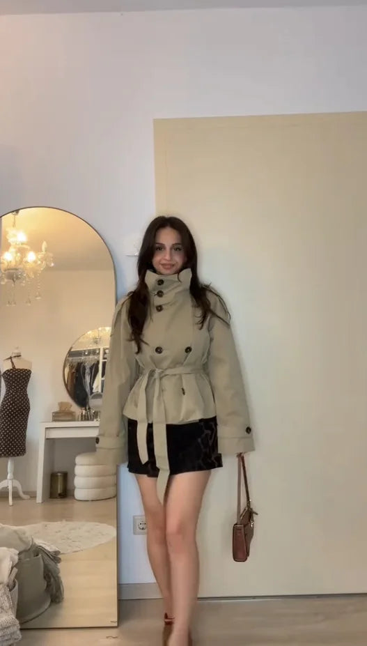Woman in belted beige trench coat and black mini skirt, standing in stylish modern room