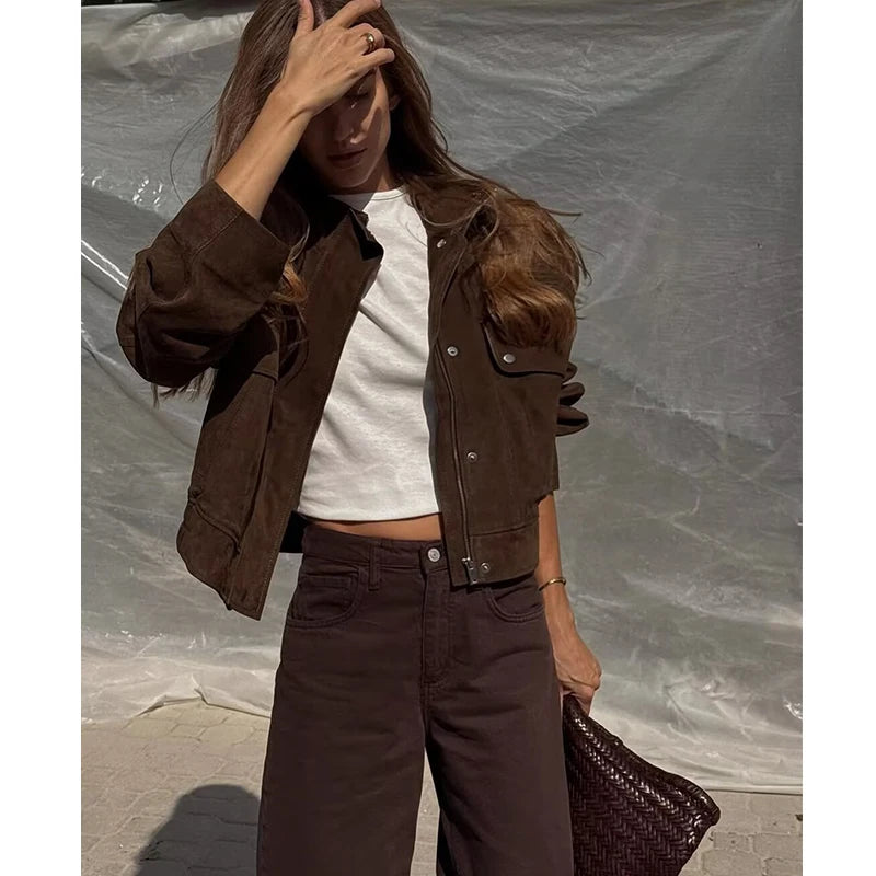 Woman wearing a brown suede jacket, white crop top, brown pants, holding woven clutch