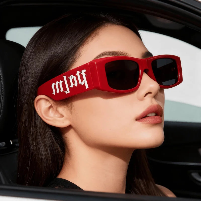Woman wearing bold red oversized sunglasses with white gothic lettering, seated in a car