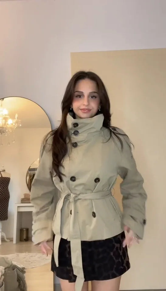Woman in a belted beige trench coat and animal print skirt standing indoors
