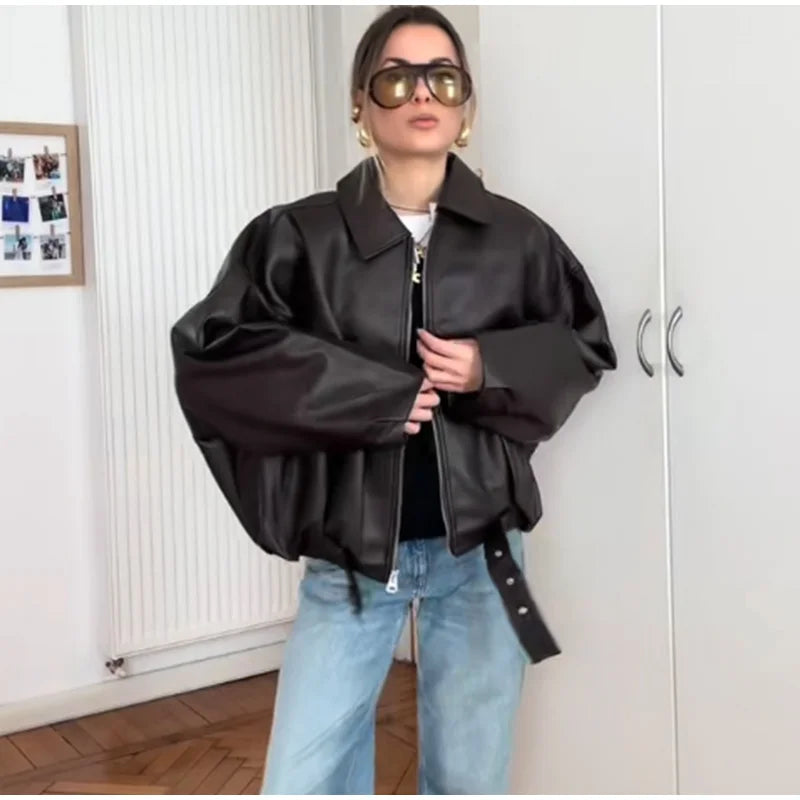 Woman in oversized black leather jacket, sunglasses, and jeans in a modern indoor setting