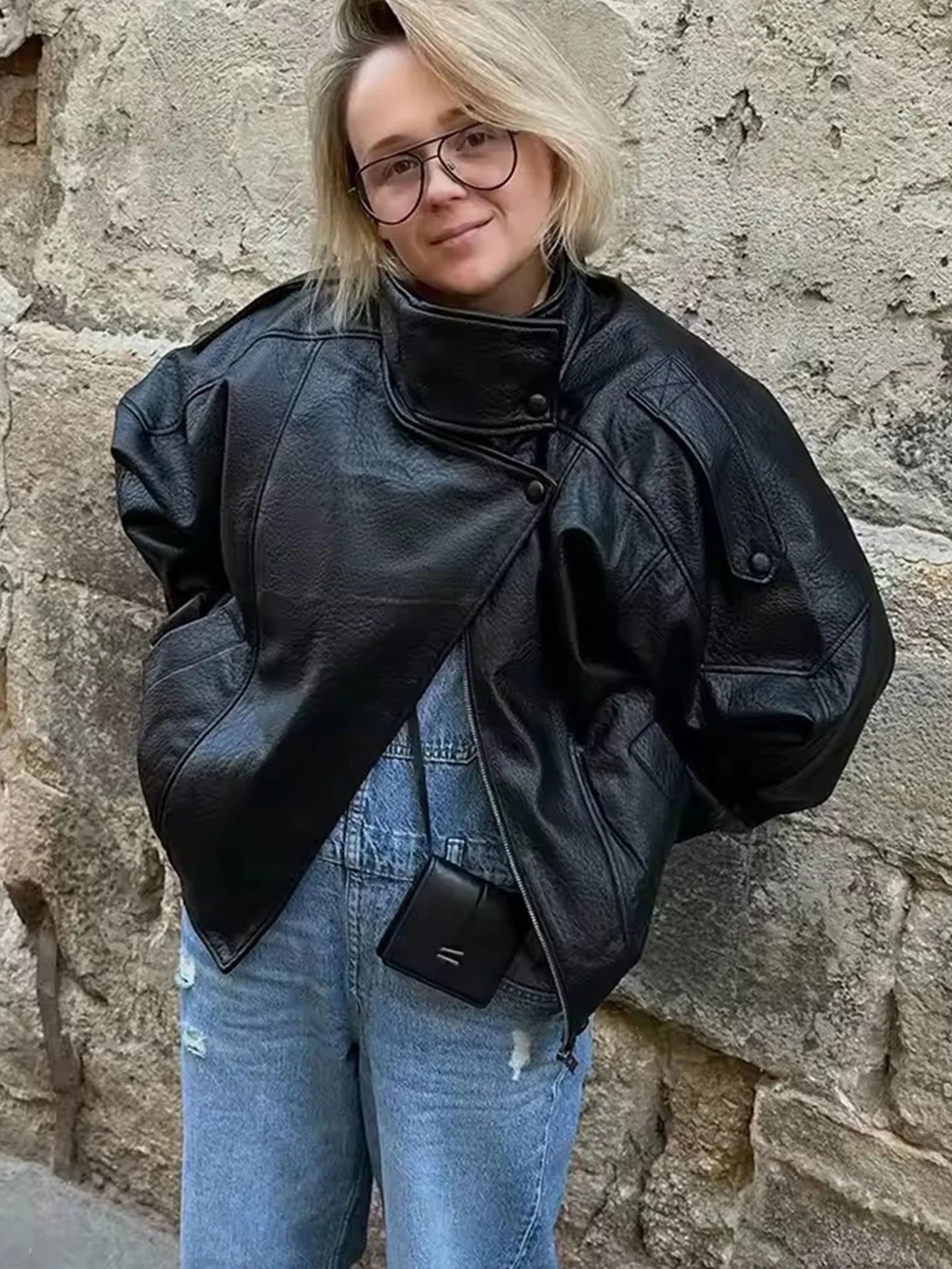 Woman in oversized black leather jacket and blue jeans with mini black bag, standing by stone wall