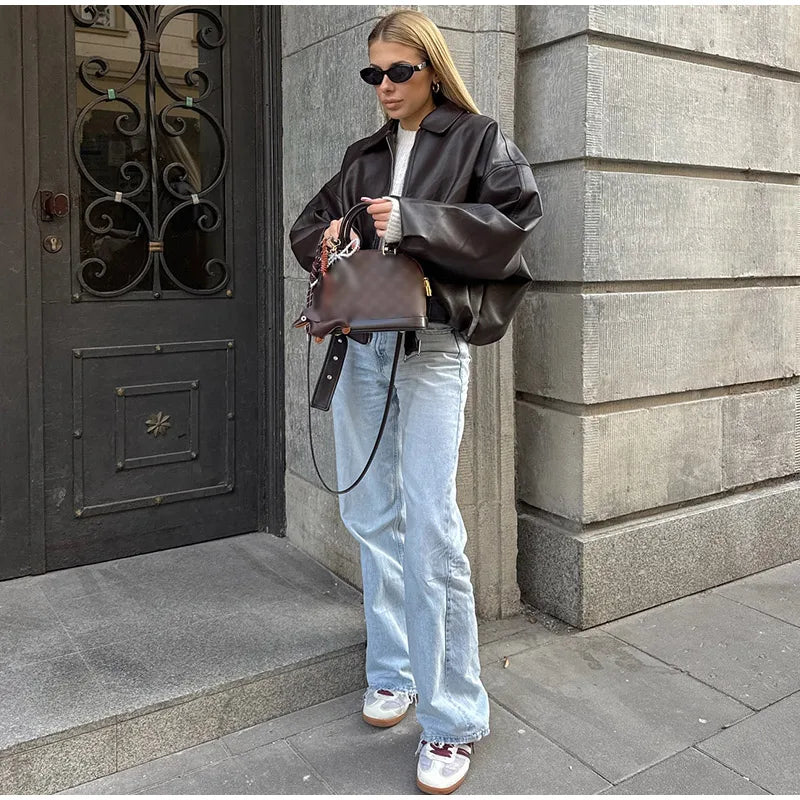 Stylish woman in oversized leather jacket, light blue jeans, sunglasses, and sneakers holding a designer handbag on city street