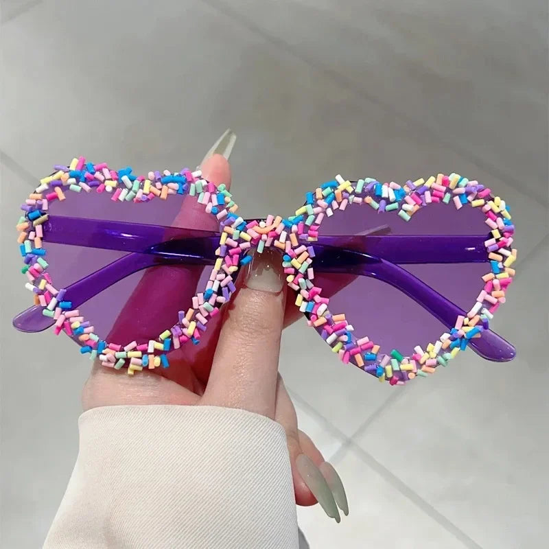Purple heart-shaped sunglasses with rainbow sprinkle accents, held by a hand indoors